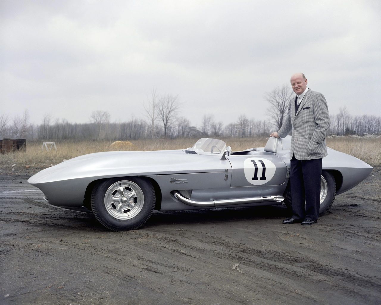 This image shows GM design chief Bill Mitchell standing proudly beside the legendary XP-87 Stingray Racer, the privately funded prototype that directly influenced the styling of the 1963 Corvette Sting Ray. Finished in silver with racing roundels, side pipes, and knock-off wheels, the Stingray was both a competition car and a rolling design laboratory. Mitchell often drove it himself, and its sharp, aerodynamic lines would become the visual DNA of the second-generation Corvette. (Image courtesy of GM Media LLC.) This image shows GM design chief Bill Mitchell standing proudly beside the legendary XP-87 Stingray Racer, the privately funded prototype that directly influenced the styling of the 1963 Corvette Sting Ray. Finished in silver with racing roundels, side pipes, and knock-off wheels, the Stingray was both a competition car and a rolling design laboratory. Mitchell often drove it himself, and its sharp, aerodynamic lines would become the visual DNA of the second-generation Corvette. (Image courtesy of GM Media LLC.)