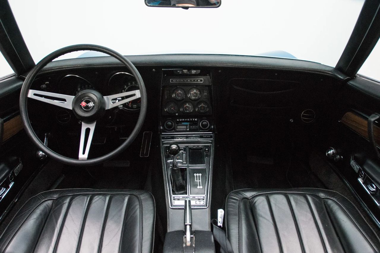 For 1970, the Corvette’s cockpit looked familiar from behind the three-spoke wheel, but the details moved upscale. High-back buckets were revised for a bit more headroom and better lateral support; the shoulder belts were tucked into the rear quarters and routed through the seatbacks; even the seat-back hinge release was easier to reach. The year’s big addition was the new “Custom Interior,” which brought leather-trimmed seat surfaces, door-sill-to-door-sill cut-pile carpet, a leather shift boot on manual cars, and wood-grain accents on the doors and console. Molded door panels with integrated armrests and storage pockets helped tidy the space, while the familiar bank of round gauges kept the look unmistakably Corvette. Subtle changes, but they collectively pushed the cabin toward a more refined grand-touring feel.