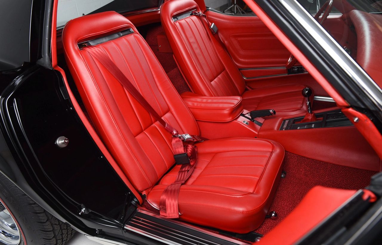 For 1970, Corvette’s cabin saw thoughtful refinements rather than a clean-sheet redo. High-back buckets introduced in ’69 were revised again, gaining about an inch of headroom and better lateral support, while the shoulder-belt inertia reels were tucked neatly into the rear quarters and the belts routed through the seatbacks for an integrated look; even the seat-back hinge release was easier to reach. The big upgrade was a new “Custom Interior,” which added leather-trimmed seat surfaces, door-sill-to-door-sill cut-pile carpet, a leather shift boot on manual cars, and wood-grain accents on the doors and console. Molded door panels with built-in armrests and storage pockets further civilized the space. Subtle changes, but they collectively made the Stingray feel more like a purposeful grand tourer inside.