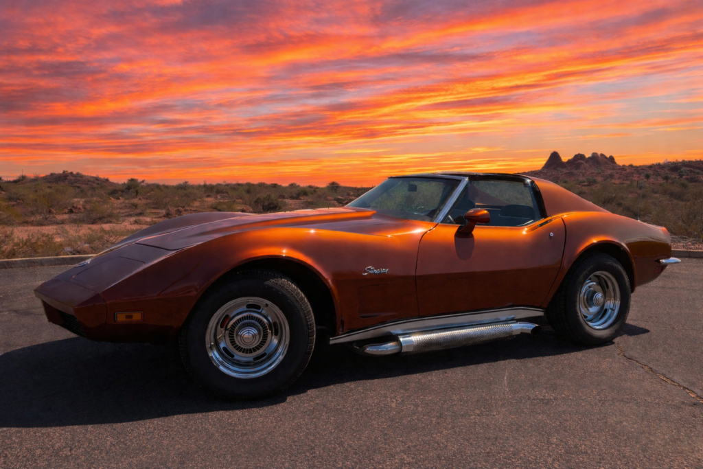 A metallic burnt-orange 1973 Chevrolet Corvette Stingray coupe is shown in side profile, parked on a paved desert turnout in Southern Arizona. The car features a long sculpted hood, removable T-top roof panels, chrome rear bumper, turbine-style wheels, and polished side-exit exhausts, all illuminated by a vivid sunset sky filled with layered orange and pink clouds. Sparse desert vegetation, saguaros, and distant rock formations stretch across the background, emphasizing the Corvette’s low stance and dramatic C3 silhouette against the open desert landscape.