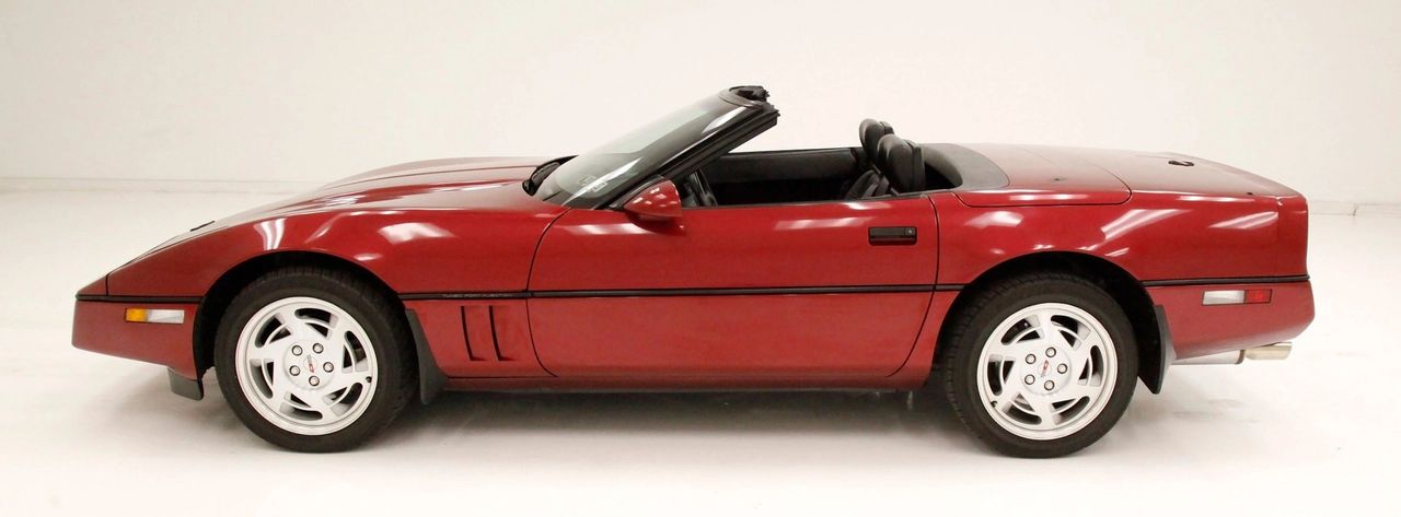 A 1990 Chevrolet Corvette Convertible, finished in Dark Red Metallic (74U), sits low and sleek with its signature wedge-shaped C4 profile. The body lines are sharp and aerodynamic, flowing from the pointed nose with its pop-up headlights to the smooth, integrated rear deck. The convertible’s soft top is neatly stowed, emphasizing the clean beltline and wide stance. The car rides on 17-inch turbine-style alloy wheels wrapped in Goodyear Eagle tires, a defining look of late-’80s and early-’90s Corvettes. Inside, the cabin features the updated 1990 dashboard with its hybrid digital/analog instrumentation and newly added passenger glovebox. The overall impression is both elegant and purposeful — a car equally at home cruising with the top down or flexing its performance heritage.