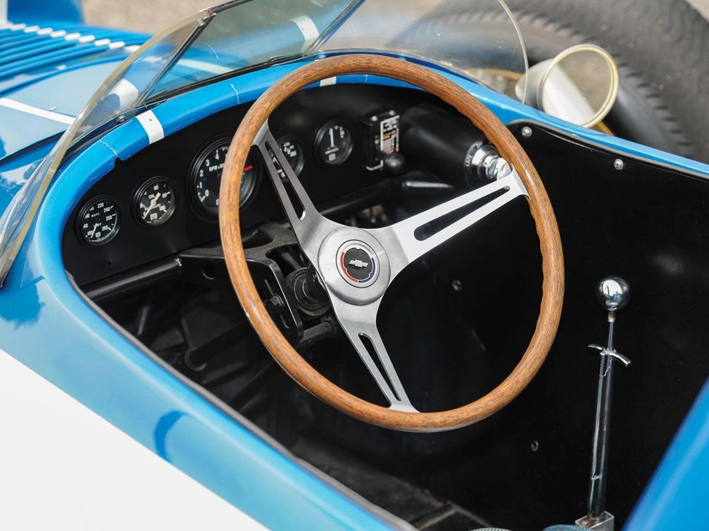 A plain, wood-rim three-spoke wheel sat at the heart of CERV I’s feedback loop—no assist, no filters, just geometry and metal. The quick 12:1 ratio (2.3 turns lock-to-lock) meant tiny inputs produced real front-wheel angle, letting the driver trim a line or catch a slide instantly. Forward-mounted, low-compliance linkages shortened the load path and kept lash out of the system, while generous caster and a small scrub radius built honest self-aligning torque without kickback. The result was high effort at walking pace but wonderfully alive at speed: surface texture, grip build, and the first hint of push or bite all arrived through the rim in real time.