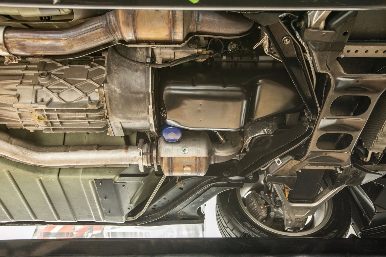 The 4L60E is on the left side of the photo—the large ribbed cast-aluminum housing just ahead of the catalytic converter and behind the exhaust Y-pipe. Compared with the earlier 4L60/700R4, the 1994 4L60E uses full electronic control of shift timing and line pressure via the PCM (shift solenoids A/B and an EPC solenoid) instead of a throttle-valve cable and hydraulic governor. Gear ratios and basic hardware stayed similar, but the “E” added computer diagnostics and adaptive shift capability, along with the necessary wiring and calibration changes. (Image courtesy of RK Motors)