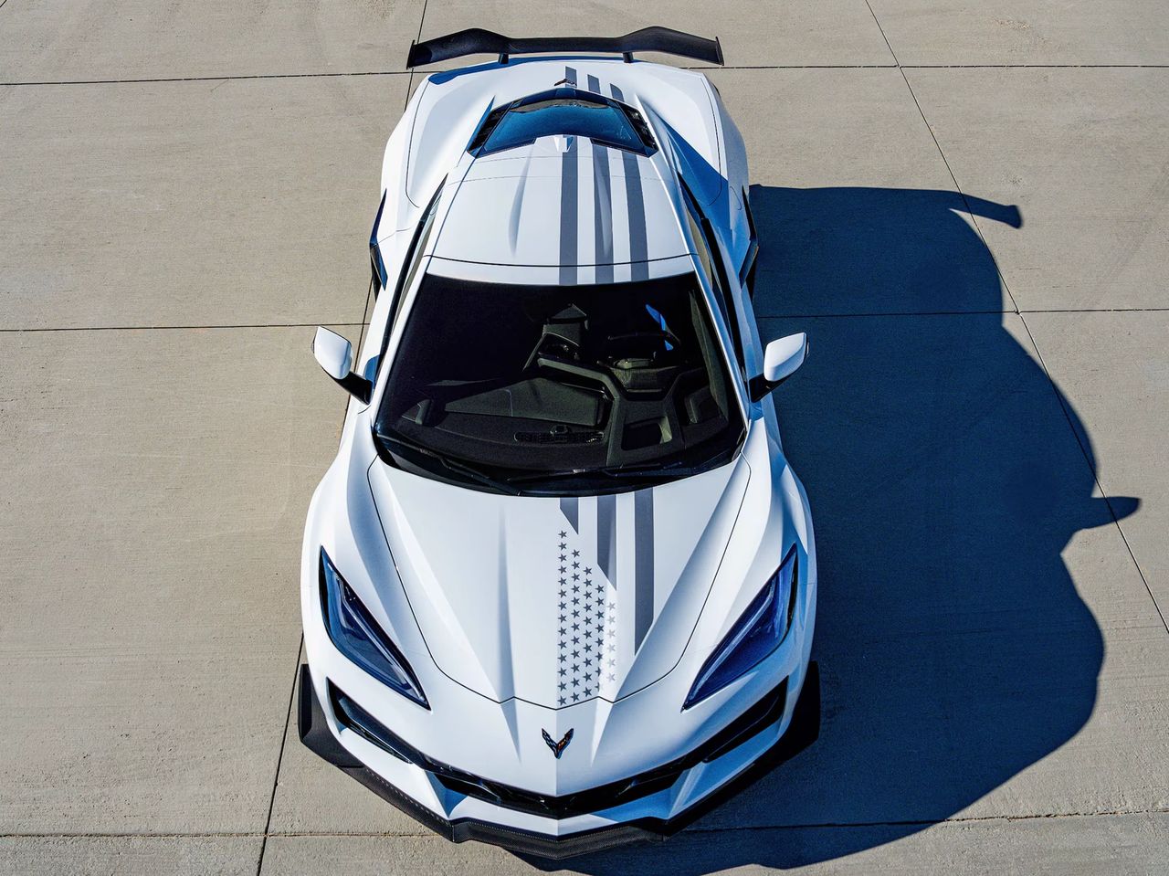 This overhead view captures the Stars & Steel theme exactly as Chevrolet intended: a clean Arctic White base punctuated by flag-inspired striping that runs nose-to-tail in a muted, metallic-looking gray (the “steel” element of the design). Front and center on the hood, the graphic transitions into a field of stars arranged as a stylized flag motif—subtle at a distance, unmistakably patriotic up close. The look is finished with the Corvette crossed-flags emblem on the nose, anchoring the package in brand identity while the Stars & Steel graphics do the commemorative heavy lifting. The overall effect is deliberate and modern—patriotism expressed through precision paintwork and restrained, premium finishes rather than loud color. (Source: GM Media)