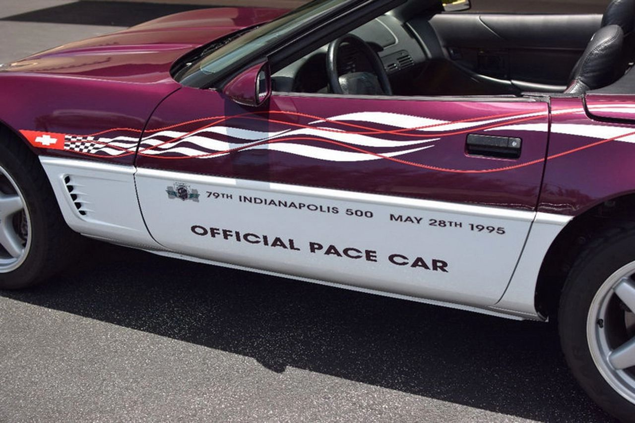 Lettered like a pit-lane credential, the Z4Z’s flanks tell the whole story: “79th Indianapolis 500 — May 28th, 1995” riding above crisp “OFFICIAL PACE CAR” script. A red tracer threads through wind-swept ribbons that split Dark Purple Metallic from Arctic White, ending at the crossed-flags fender badge. The close-up also shows late-C4 cues—the clean side gills, body-color handle, and five-spoke alloys—wrapped in a livery that looks in motion even when parked.