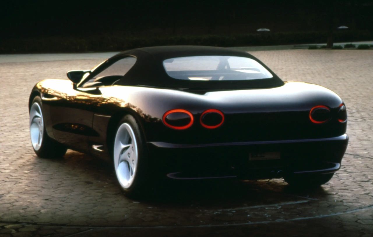 From this rear three-quarter angle, the Stingray III reveals several of its most distinctive—and forward-looking—design cues. The rounded taillamps, set into a smooth, almost liquid surface, were a deliberate evolution of Corvette’s trademark quad-light theme. Instead of being recessed or split by bodywork, they float in a clean horizontal line, previewing the flush, minimalist rear ends that would dominate GM design language in the 1990s. The sharply tucked rear fascia and integrated exhaust outlet mark a clear departure from the blockier, bumper-defined C4 Corvette, hinting at the flowing surfaces that would arrive with the C5. The roofline and expansive glass taper down gracefully, emphasizing aerodynamics and cab-rearward stance—an element that pushed Corvette closer to its exotic European rivals. Finally, the wide five-spoke wheels, pushed out to the corners, give the car a planted, muscular presence. Each of these choices underscored the Stingray III’s mission: to prove Corvette design could move beyond the digital angularity of the 1980s and embrace organic curves and restrained sophistication for the future.