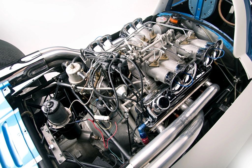 A mid-mounted Chevrolet small-block sits like a lab experiment, wearing an independent-runner intake with eight velocity stacks and Hilborn-style mechanical fuel injection—barrel valve, individual injector lines, and all—for razor response and cylinder-by-cylinder tuning. The external oil tank and scavenge plumbing flag a dry-sump system, letting the engine ride low without oil starvation, while equal-length headers sweep into polished megaphones to clear heat and let the V-8 breathe. CERV I cycled through several engines during development—starting with a Rochester-injected 283, then high-output 327s, and ultimately an all-aluminum 377-cid package around the 500-hp mark—and the eight-stack, dry-sump hardware you see here matches that later 377-cid configuration. True to CERV I’s mission, every line, fitting, and linkage is exposed for fast changes and clean data at the track; it’s a purpose-built testbed disguised as an engine bay.