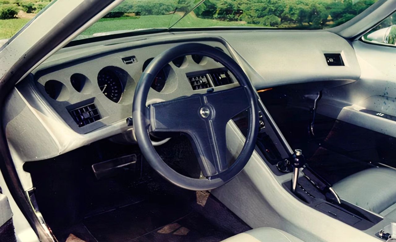 Inside the XP-987 GT, the cabin is as experimental as the car’s rotary heart, with a sweeping dash that wraps around the driver in a clean, almost architectural arc. Deep-set round gauges, simple rectangular vents, and the minimalist three-spoke wheel give the cockpit a purposeful, almost aircraft-like feel. The high, narrow console and upright shifter emphasize that this was meant to be driven, not just displayed. It’s a fascinating bridge between late C3 ergonomics and the driver-focused layouts that would later define modern Corvettes. Inside the XP-987 GT, the cabin is as experimental as the car’s rotary heart, with a sweeping dash that wraps around the driver in a clean, almost architectural arc. Deep-set round gauges, simple rectangular vents, and the minimalist three-spoke wheel give the cockpit a purposeful, almost aircraft-like feel. The high, narrow console and upright shifter emphasize that this was meant to be driven, not just displayed. It’s a fascinating bridge between late C3 ergonomics and the driver-focused layouts that would later define modern Corvettes.