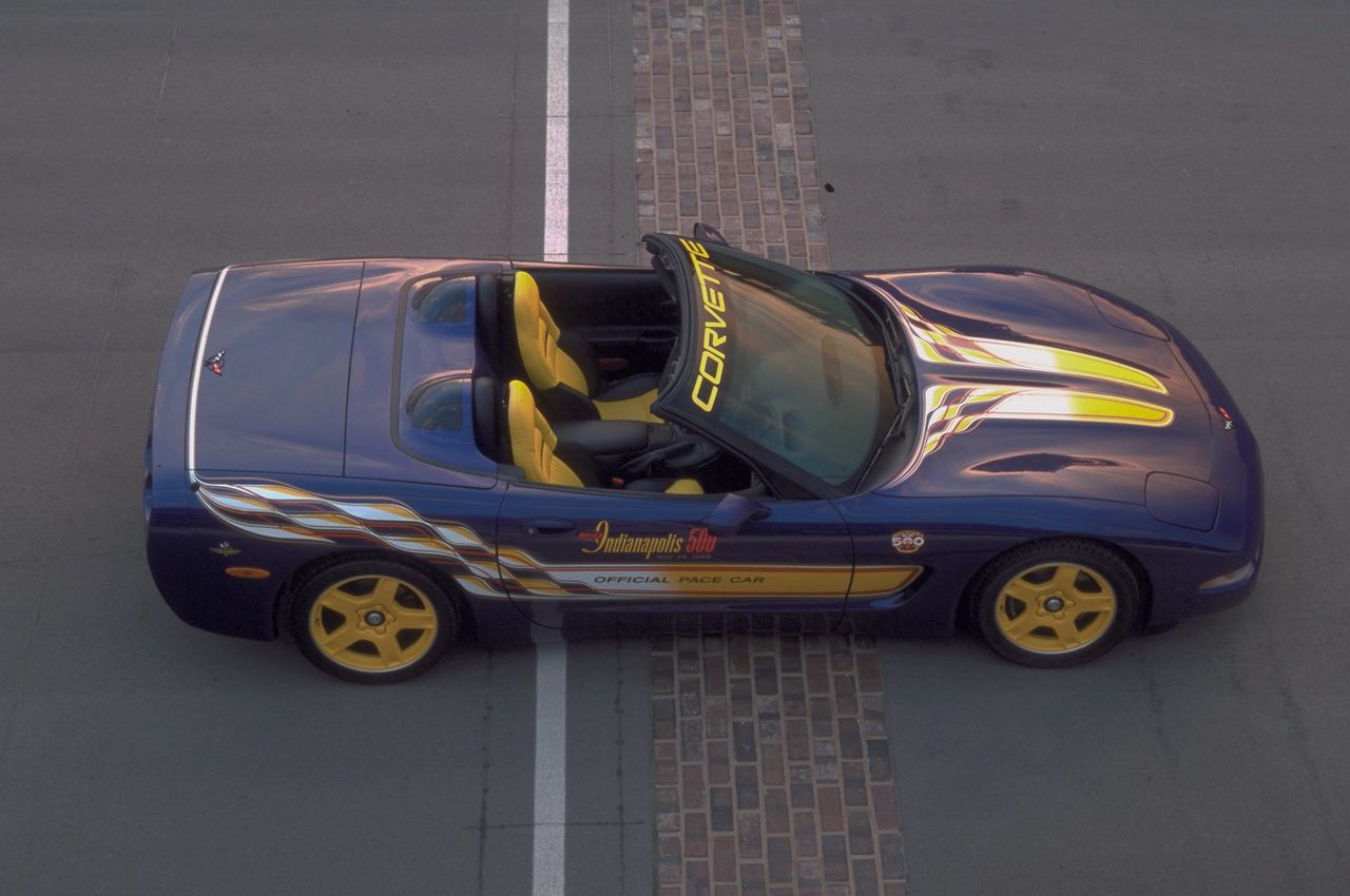 Indianapolis Motor Speedway’s iconic brick start/finish line, seen from above—the 1998 Corvette Indy 500 Pace Car straddles the stripe, Radar Blue and yellow livery laid out like a starter’s flag. The CORVETTE windshield banner, triple hood streaks, and checkerboard ribbon graphics broadcast its mission at a glance.