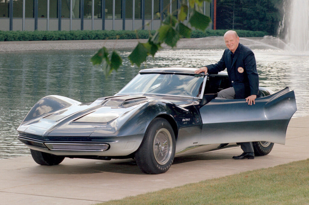 Bill Mitchell stands beside the open door of the blue-and-silver 1965 Mako Shark II near the GM Design Center fountain, highlighting the concept’s dramatic shark-inspired gradient paint.