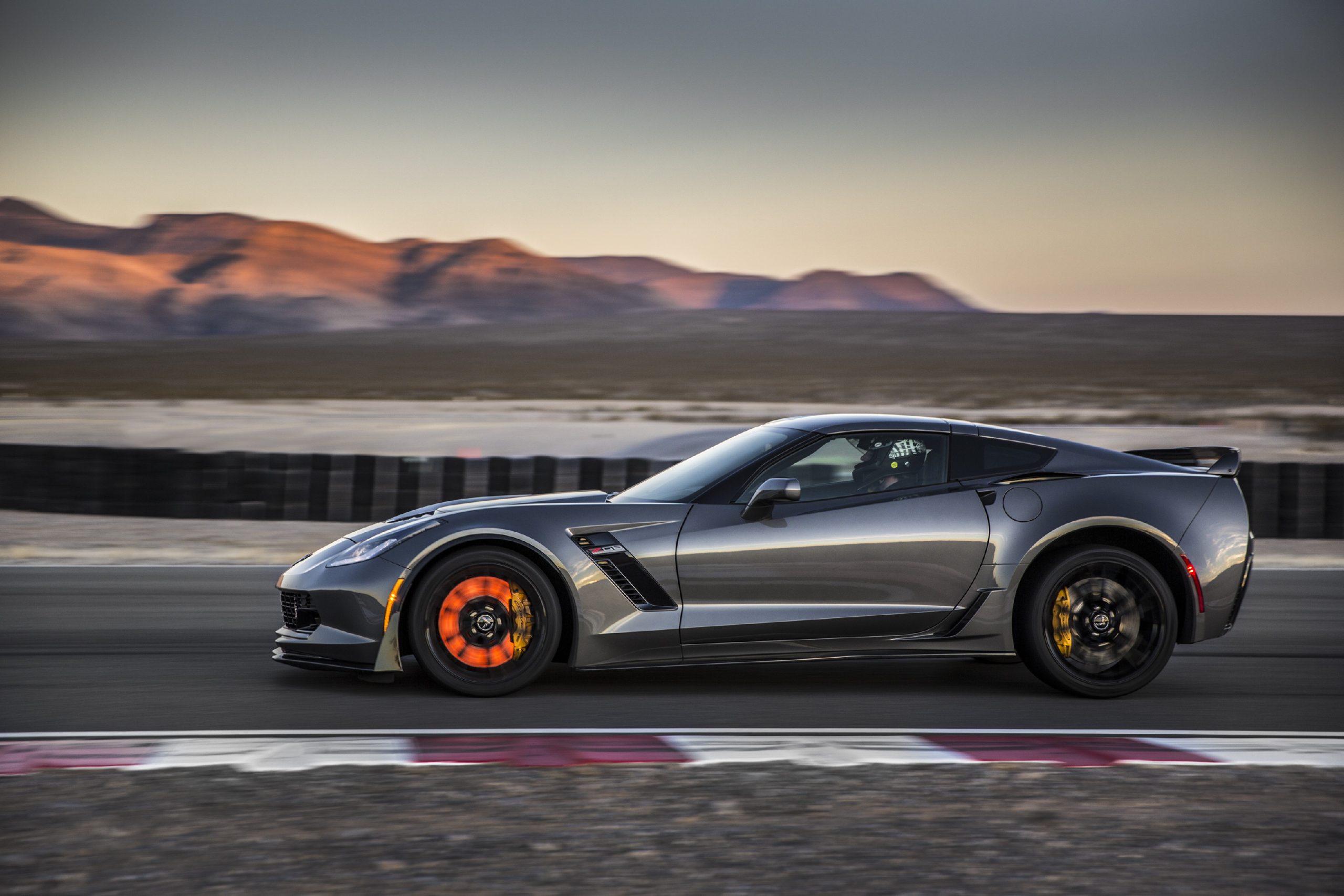 Shark Gray 2015 Chevrolet Corvette Z06 at speed on a racetrack at dusk, shown in side profile with a helmeted driver and its front brake rotor glowing bright orange under heavy braking.