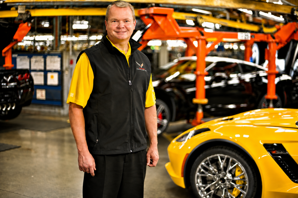 Kai Spande stands on the Bowling Green Assembly Plant production floor beside a yellow C7 Corvette during his tenure as plant manager, surrounded by Corvette manufacturing equipment and vehicles on the assembly line.