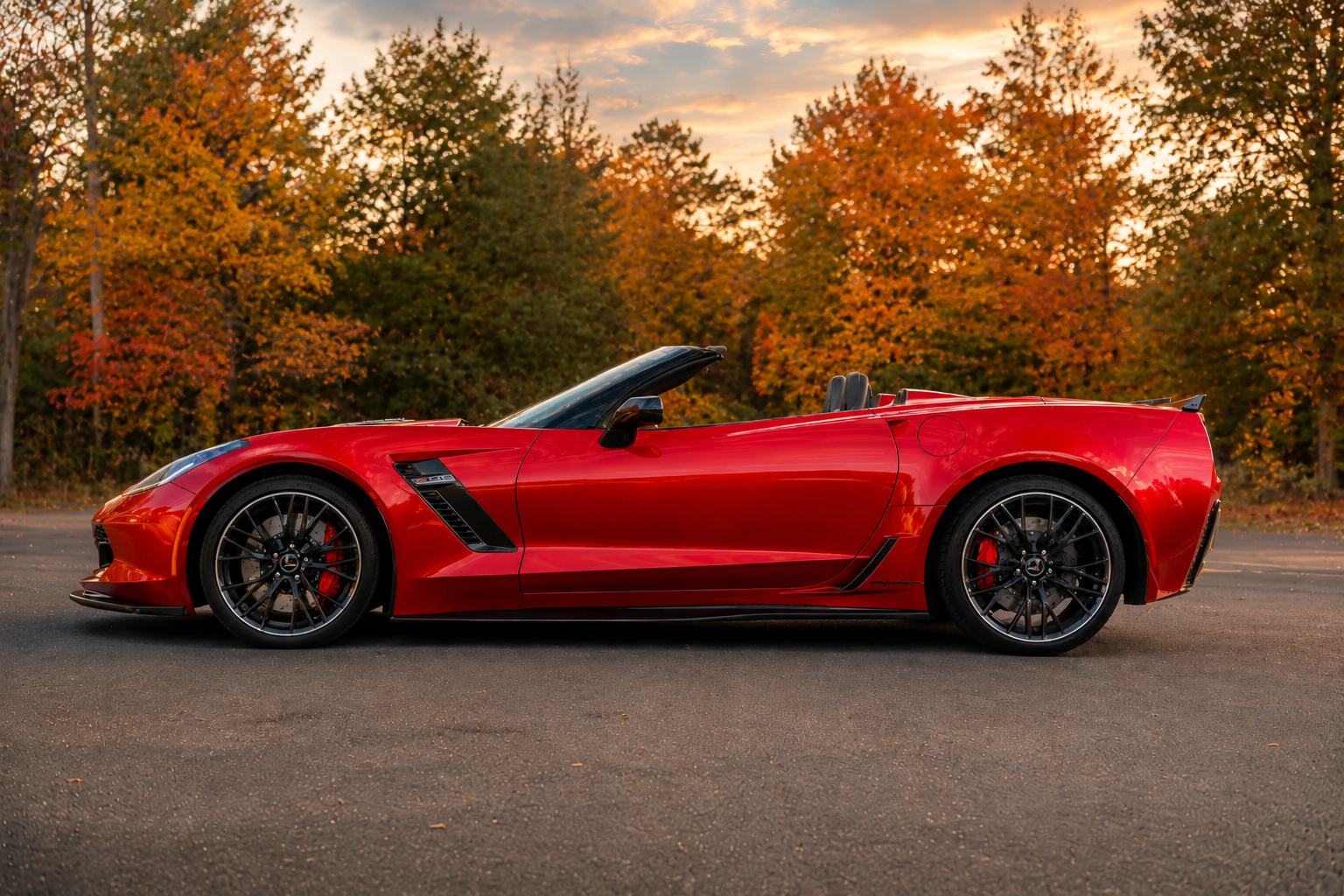 Side-profile view of a red 2015 Chevrolet Corvette Z06 Convertible with the top down, parked on pavement against a scenic autumn backdrop with colorful fall trees and a warm early-evening sky.