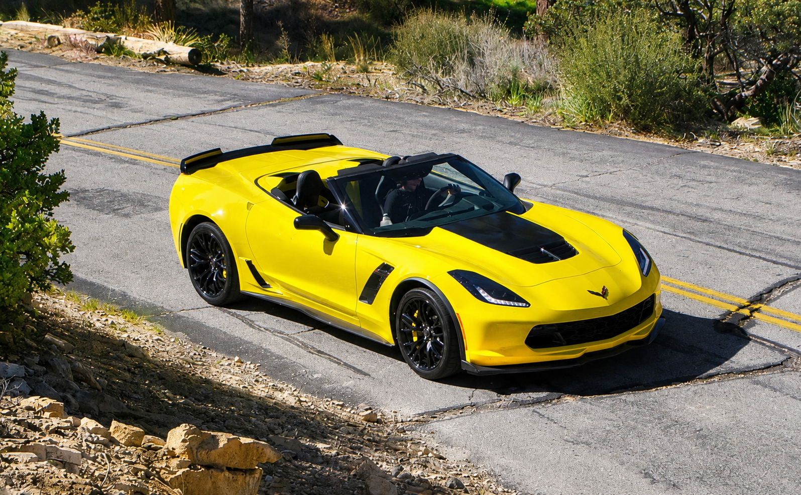 Yellow 2015 Chevrolet Corvette Z06 Convertible with the top down, photographed from an elevated front three-quarter angle while driving along a winding two-lane road.