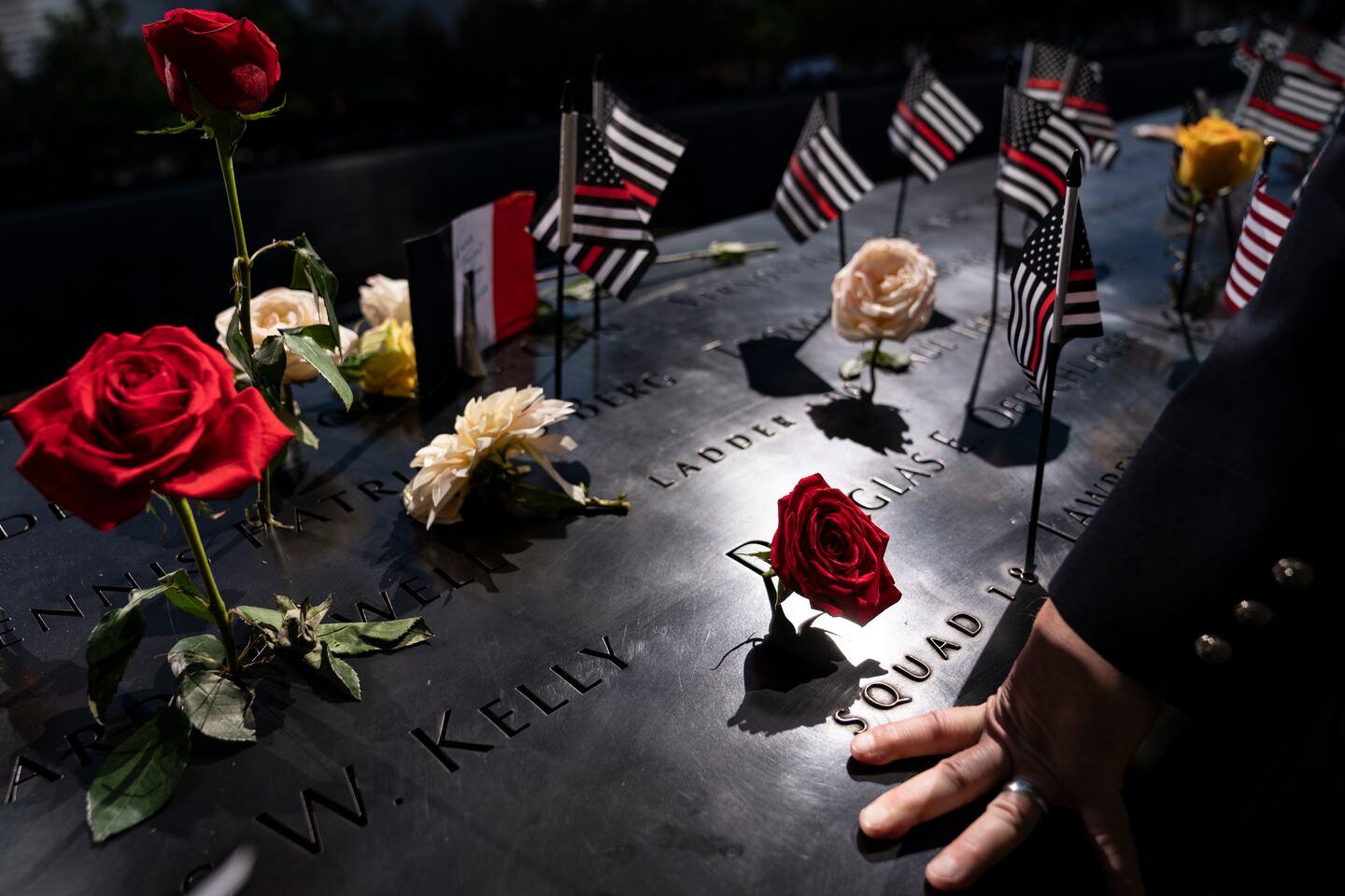 Flowers and memorial flags surround engraved names at the National September 11 Memorial as a visitor rests a hand against the bronze parapet in a quiet act of remembrance.
