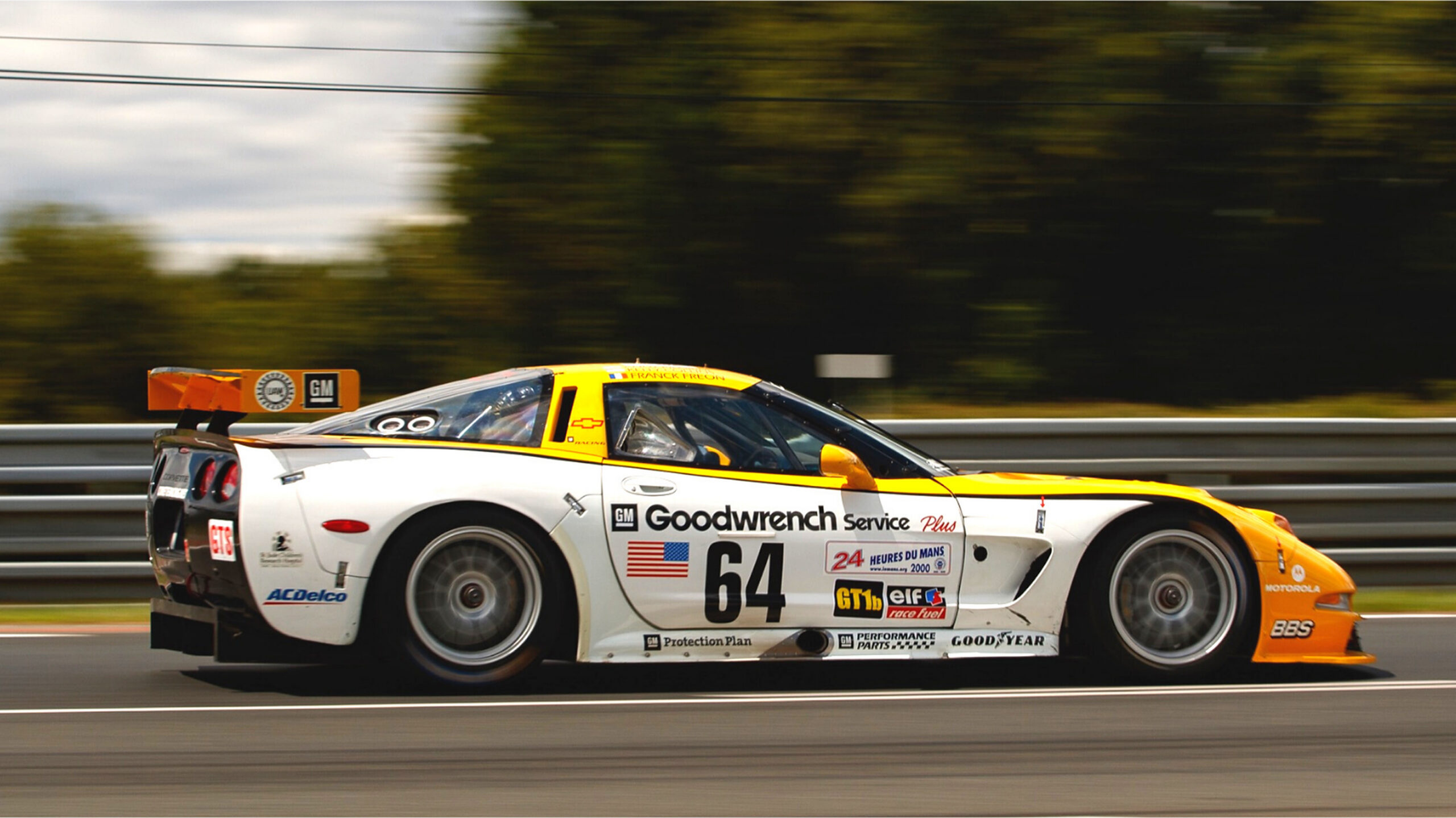 Side-profile action photograph of the No. 64 Chevrolet Corvette C5-R competing at the 2000 24 Hours of Le Mans, featuring its distinctive white, yellow, and black GM Goodwrench Service Plus racing livery, Chevrolet and GM sponsorship decals, large rear wing, and Goodyear racing tires. The Corvette is captured at speed along the circuit with a motion-blurred background.