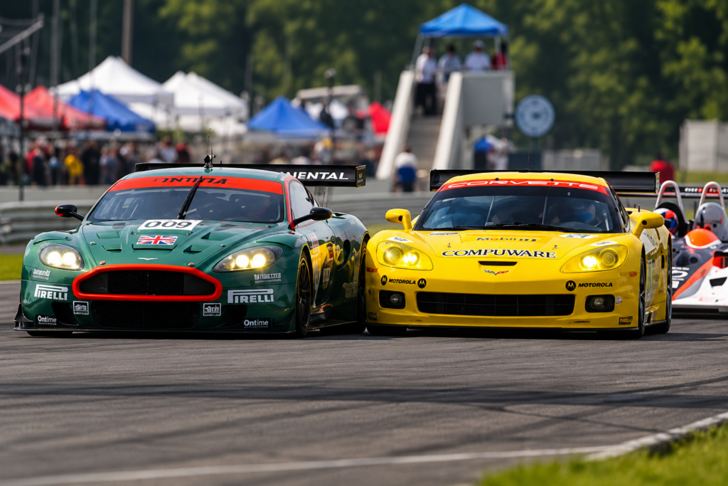 Dramatic head-on racing photograph from the 2006 American Le Mans Series showing a British Racing Green Aston Martin DBR9 and a bright yellow Chevrolet Corvette C6.R competing side by side on the racetrack, their headlights illuminated as they battle for position. A prototype race car follows closely behind, with spectators, racing paddock tents, and trackside structures visible in the background.