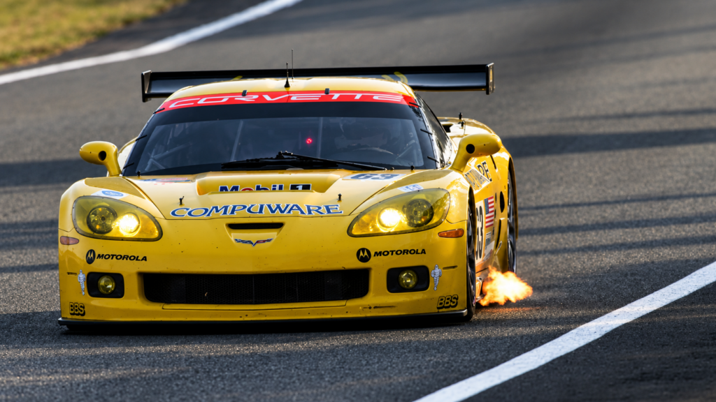 Front-facing action photograph of a yellow Chevrolet Corvette C6.R GT1 race car competing at the 2006 24 Hours of Le Mans, featuring illuminated headlights, Compuware and Mobil 1 sponsorship graphics, a large black rear wing, and a bright orange flame erupting from the passenger-side exhaust as the Corvette accelerates along the circuit.