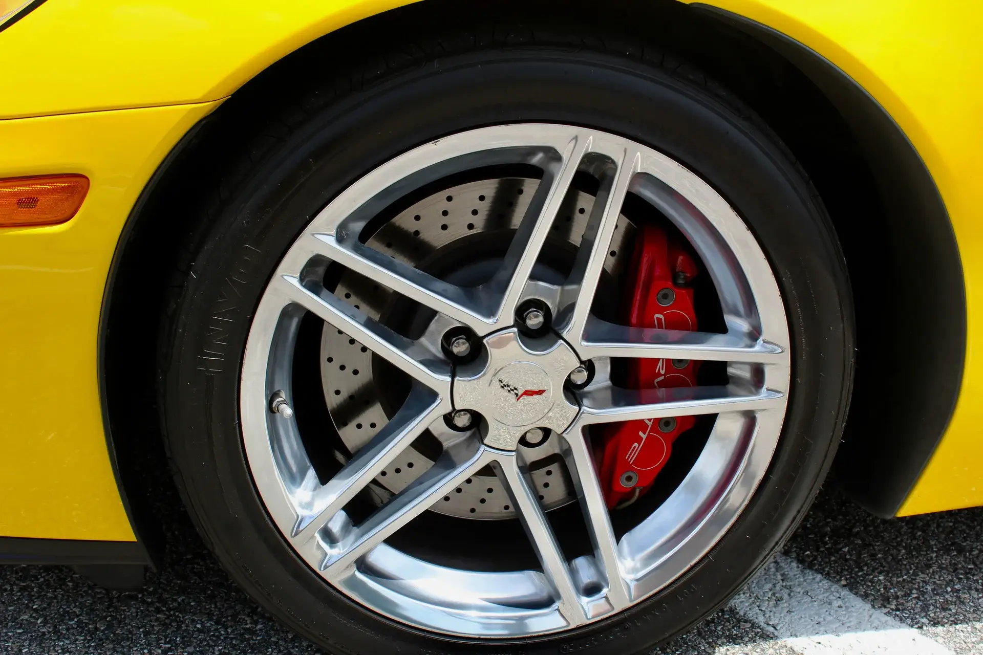 Close-up of the front wheel and upgraded braking system on a yellow 2006 Chevrolet Corvette Z06, featuring a polished aluminum split-spoke wheel, large cross-drilled brake rotor, and distinctive red six-piston Z06 brake caliper.