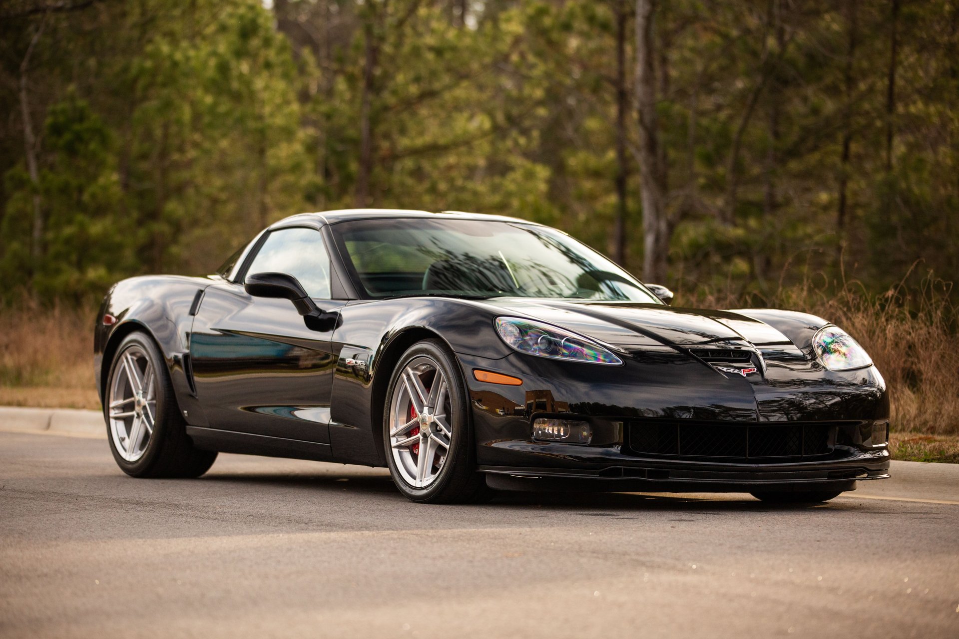 Front three-quarter view of a black 2006 Chevrolet Corvette Z06 (C6) coupe parked on a paved road against a wooded backdrop. The photograph highlights the Z06’s aggressive front fascia, distinctive raised hood with functional air intake, widened fenders, exposed projector-style headlamps, front brake cooling ducts, and polished five-spoke alloy wheels with red brake calipers. The Corvette’s glossy black finish accentuates its sculpted bodywork, low stance, and aerodynamic design, showcasing the distinctive styling of Chevrolet’s 505-horsepower, LS7-powered performance flagship.