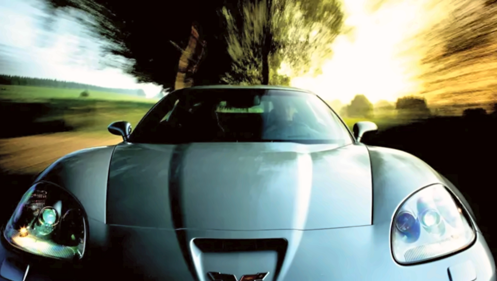 Silver 2006 Chevrolet Corvette Z06 photographed head-on while traveling along a tree-lined road, with sunlight filtering through the surrounding landscape. The dramatic low-angle photograph highlights the Z06's sculpted hood, central cooling opening, exposed projector headlights, and wide front bodywork, while the motion-blurred background emphasizes speed and performance.