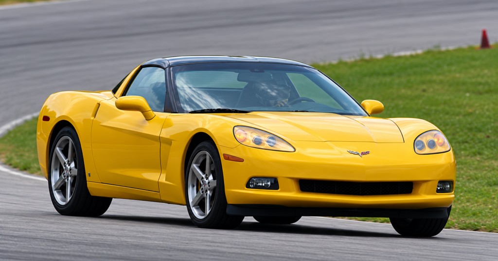 A yellow 2006 Chevrolet Corvette C6 coupe accelerating through a sweeping corner on a racetrack, photographed from a front three-quarter angle. The car features a removable black roof panel, exposed projector-style headlights, five-spoke alloy wheels, sculpted bodywork, and the Corvette crossed-flags emblem. A helmeted driver is visible through the windshield, while the paved racing surface, grassy infield, and distant traffic cone provide the backdrop for this demonstration of the 400-horsepower LS2-powered Corvette coupe’s performance capabilities.