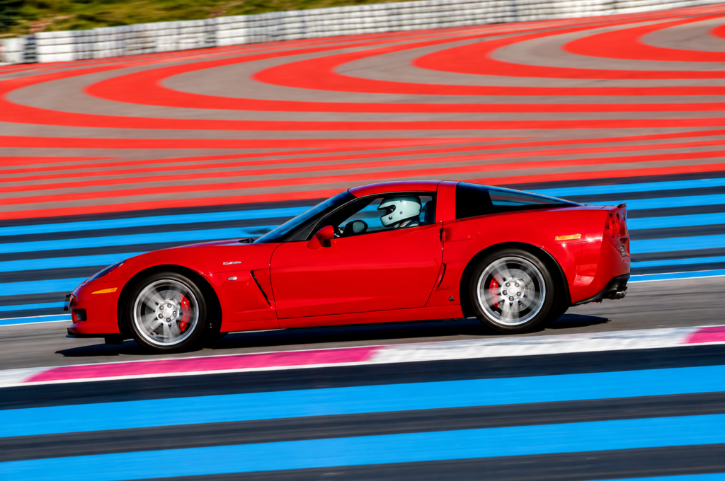 Red 2006 Chevrolet Corvette Z06 accelerating around a racetrack, photographed in profile with a helmeted driver visible through the side window. The image highlights the car's low stance, front aerodynamic splitter, sculpted side bodywork, wide rear fenders, and integrated rear spoiler.