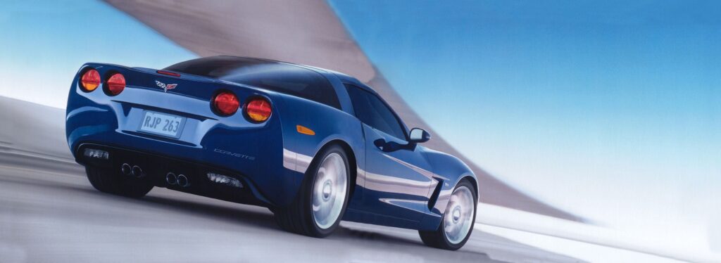 Rear three-quarter action view of a blue 2006 Chevrolet Corvette C6 coupe driving along a sweeping roadway. The image highlights the Corvette’s four round taillamps, centered quad exhaust outlets, sculpted rear fascia, distinctive side coves, five-spoke alloy wheels, and sloping rear glass. Motion blur in the wheels and pavement emphasizes the car’s speed, while the dramatic blue-and-silver background complements its metallic paint finish. The photograph captures the sixth-generation Corvette’s timeless styling and performance character, illustrating its growing appeal as a modern collector car.