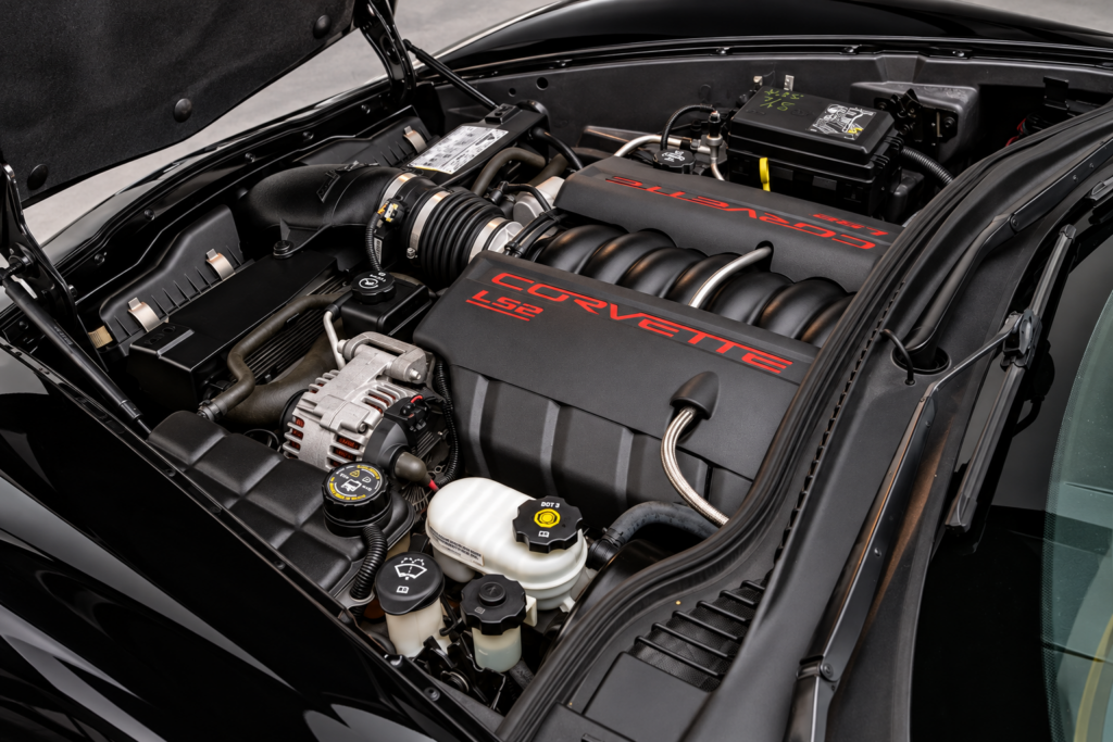 Close-up view of the 6.0-liter LS2 V8 engine installed in a black 2006 Chevrolet Corvette C6, showing the black engine covers with red Corvette and LS2 lettering, intake manifold, alternator, air intake assembly, fluid reservoirs, and surrounding engine bay components. The photograph highlights the clean, compact layout of Chevrolet’s 400-horsepower aluminum small-block V8.