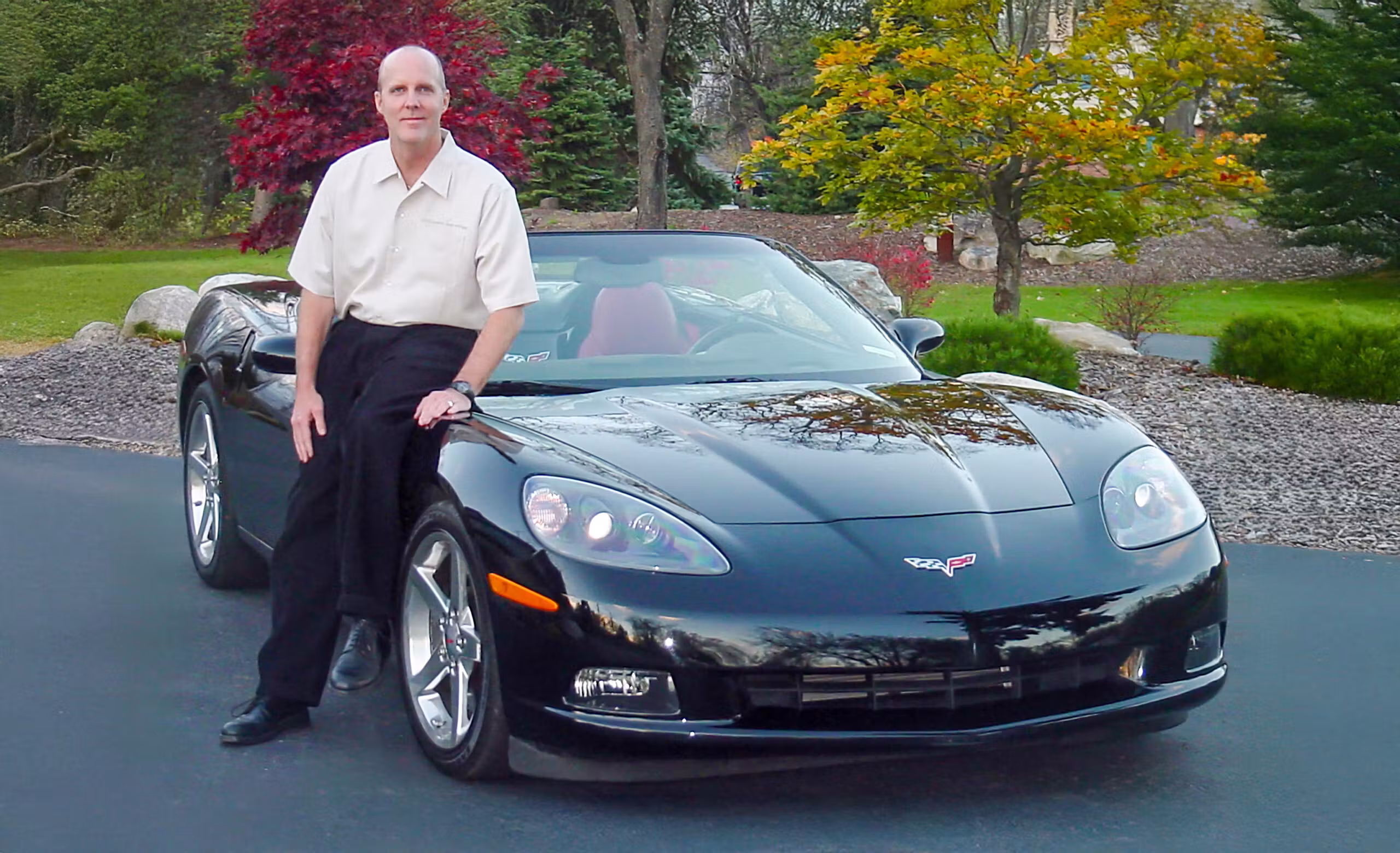 Corvette chief designer Tom Peters seated on the front fender of a black sixth-generation Chevrolet Corvette convertible with a red interior, photographed outdoors against a landscaped backdrop of green grass, trees, and autumn foliage. Peters led the design of the C6 Corvette, introduced for the 2005 model year, and its styling evolution into the 2006 Z06.