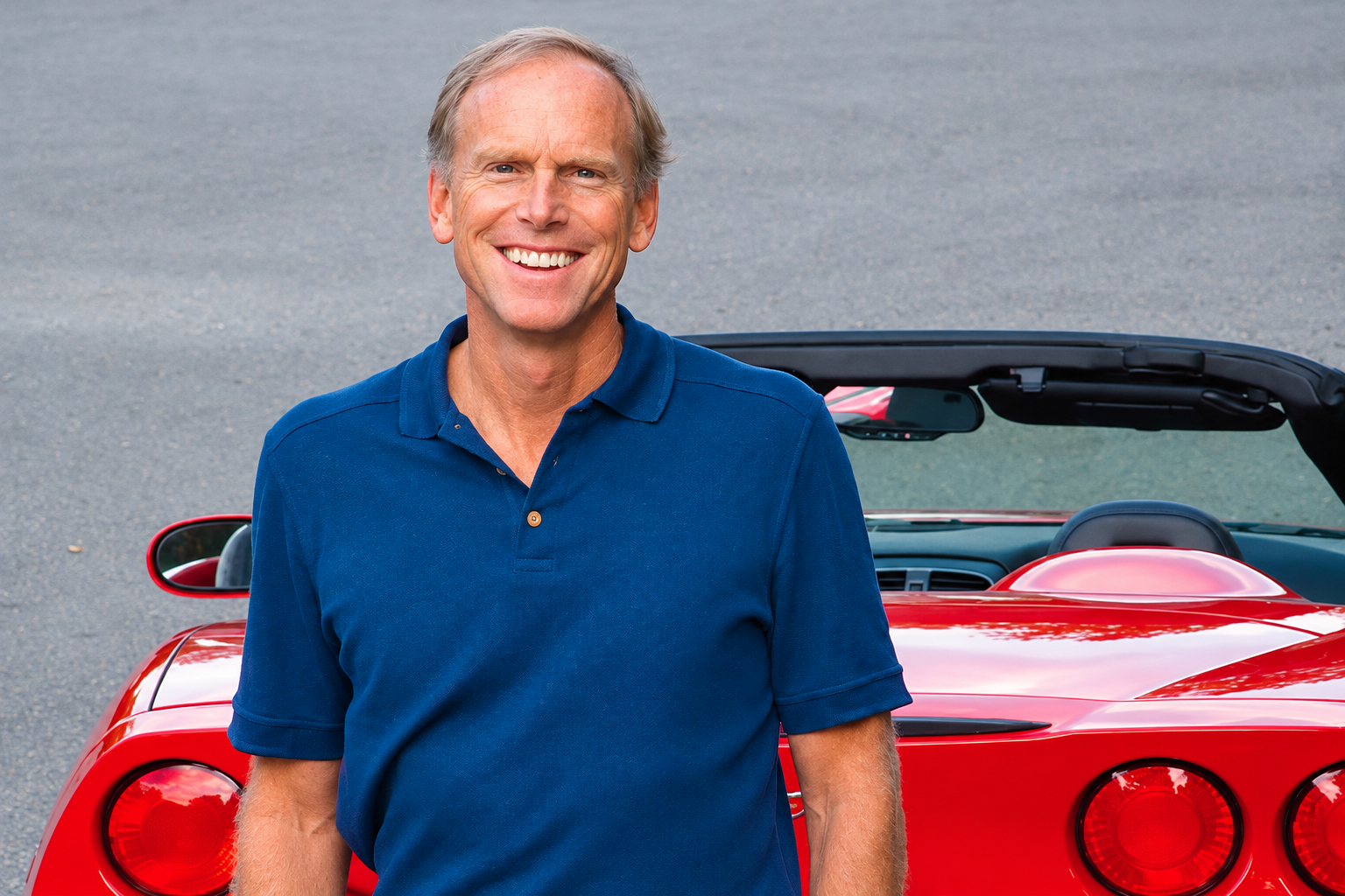 Corvette chief engineer Tadge Juechter, wearing a blue polo shirt, stands smiling in front of a red Chevrolet Corvette convertible with its top lowered. Juechter became Corvette's fifth chief engineer in July 2006, following significant involvement in the development of the C5 and C6 generations.