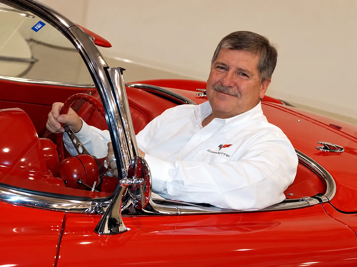 Tom Wallace, Corvette's fourth chief engineer and former Vehicle Line Executive for GM Performance Cars, seated behind the wheel of a classic red Chevrolet Corvette convertible. Wallace wears a white Corvette-branded shirt and sits in the car's red interior, surrounded by chrome windshield trim and vintage Corvette styling details.