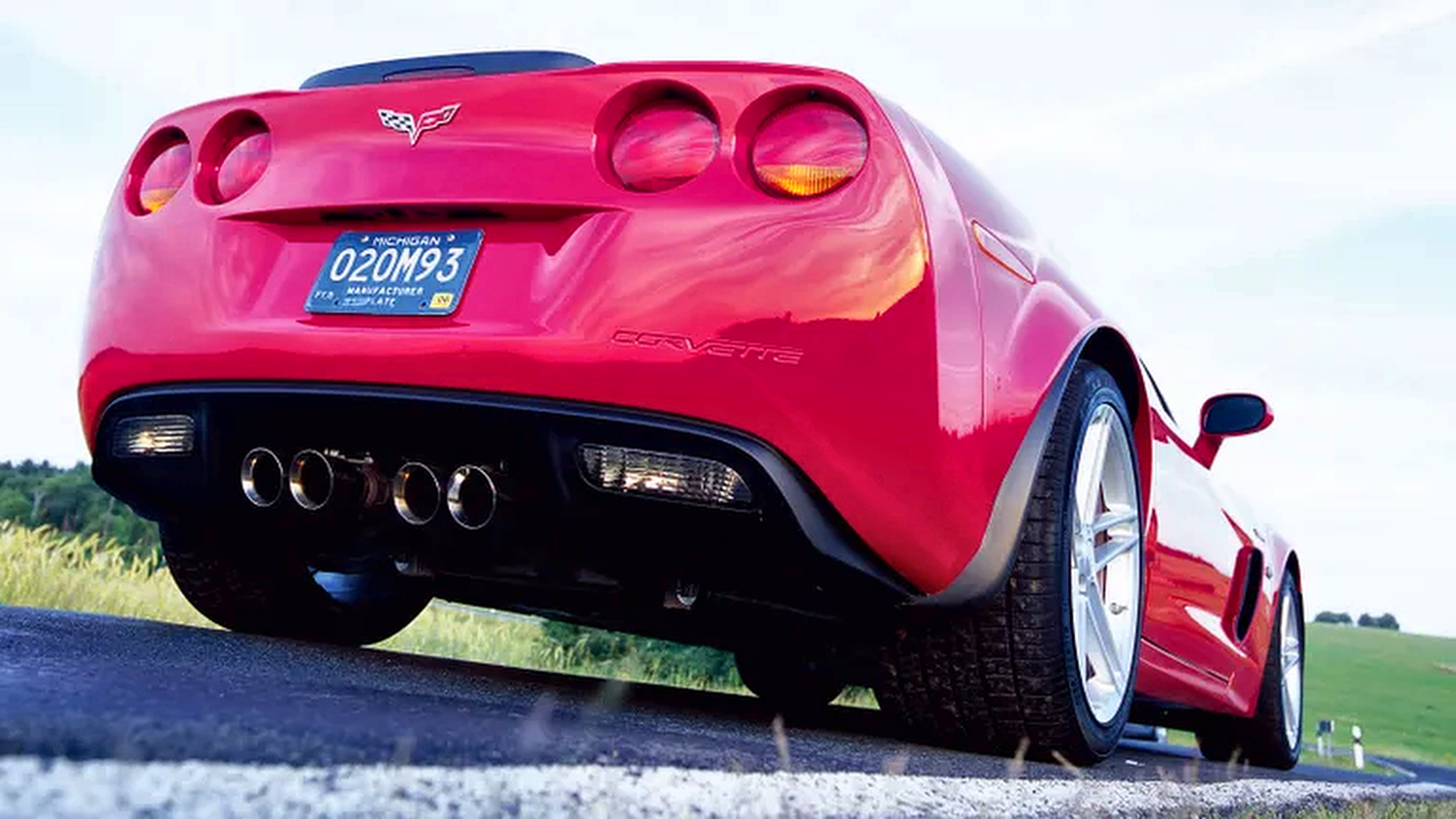 Rear three-quarter, low-angle view of a red 2006 Chevrolet Corvette C6 Z06 parked on a paved road, showcasing its four center-mounted exhaust outlets, distinctive rear fascia, wide performance tires, and silver alloy wheels against a rural landscape.