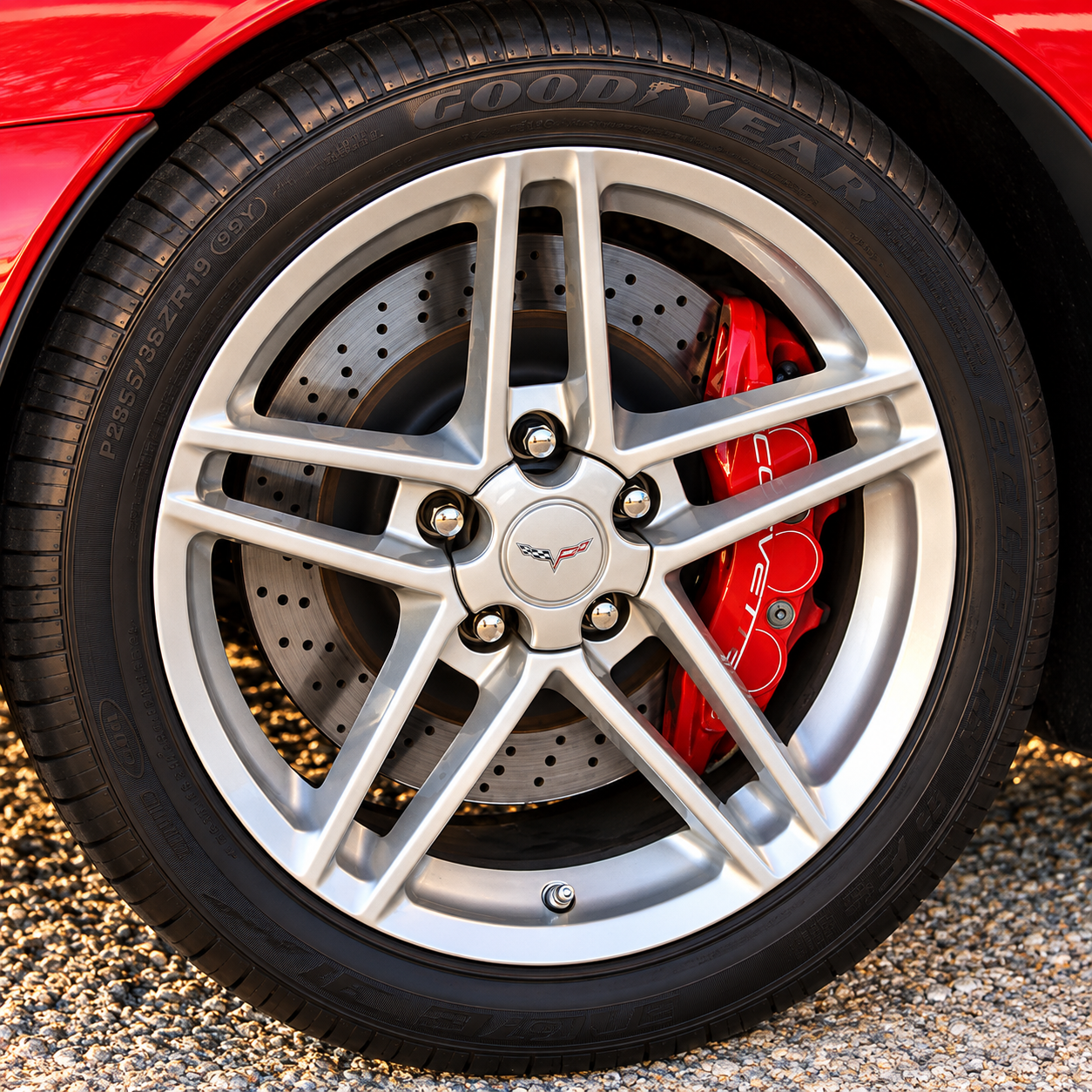 Close-up of a 2006 Chevrolet Corvette Z06 wheel and braking assembly, highlighting the silver split-spoke aluminum wheel, large cross-drilled and ventilated brake rotor, and red Corvette-branded brake caliper. The photograph illustrates the high-performance braking hardware developed to deliver consistent stopping power and resist brake fade during repeated high-speed driving.