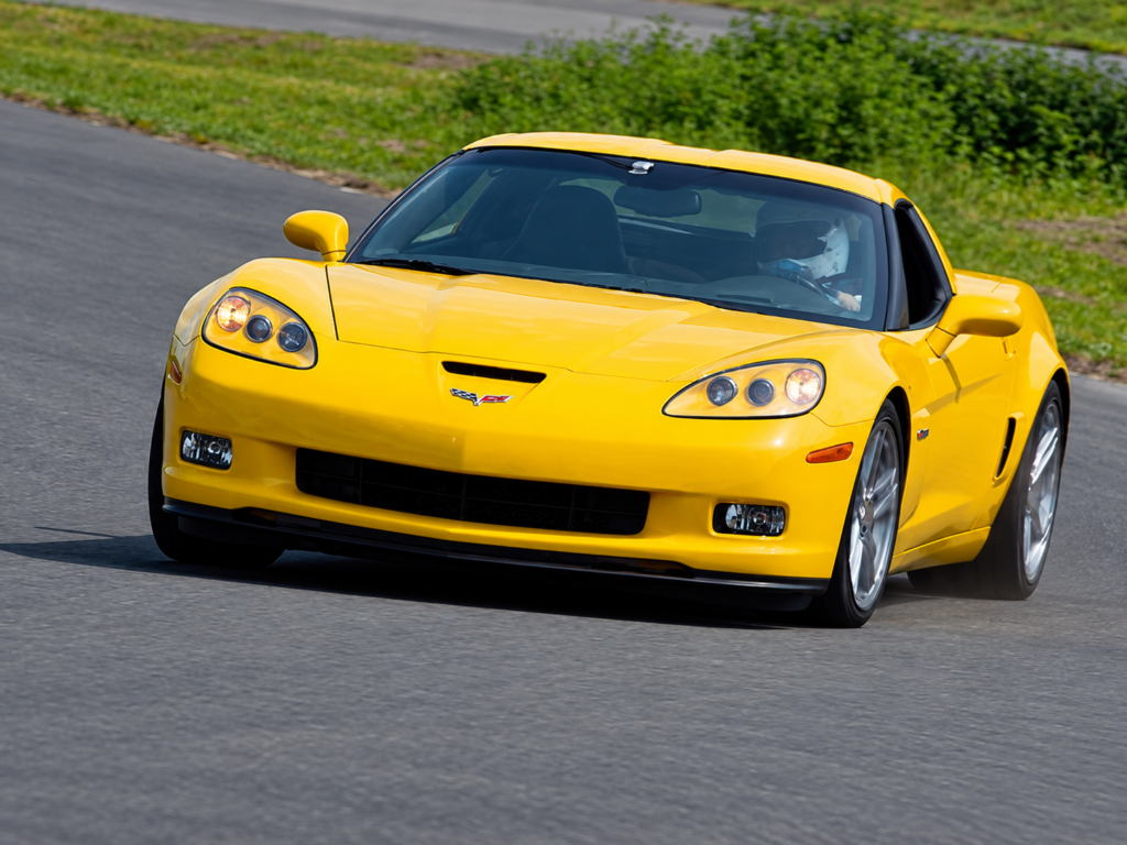 Yellow 2006 Chevrolet Corvette Z06 negotiating a sweeping corner on a racetrack, photographed from a front three-quarter angle. The image highlights the Corvette's wide stance, low aerodynamic profile, performance tires, and controlled body movement during high-speed cornering, with a helmeted driver visible through the windshield.