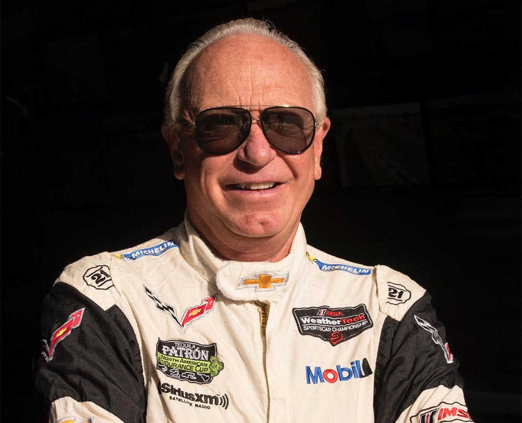 Portrait of Doug Fehan, longtime Corvette Racing Program Manager, smiling while wearing dark sunglasses and a white-and-black Chevrolet Corvette Racing suit featuring Chevrolet, Michelin, Mobil 1, and IMSA WeatherTech SportsCar Championship insignia.