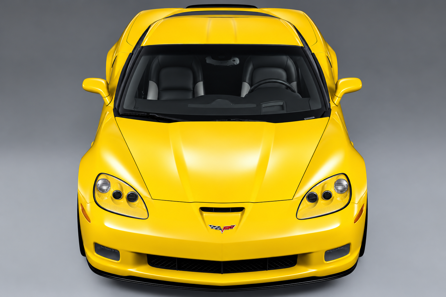 High-resolution, elevated front view of a Velocity Yellow 2006 Chevrolet Corvette Z06 (C6) against a neutral gray studio background. The photograph highlights the Z06’s distinctive raised hood with functional air intake, widened front fenders, exposed projector-style headlamps, aggressive front fascia, lower grille opening, and black front splitter. The Corvette crossed-flags emblem is prominently displayed on the nose, while the windshield reveals the black sport seats and driver-focused cockpit.